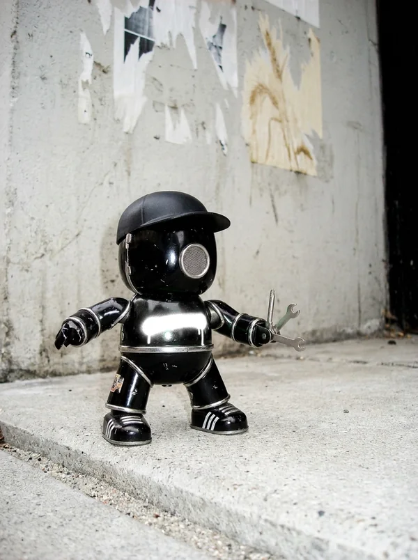 Kleiner schwarzer Blechroboter mit Cap, Lautsprechermund und Taschenmesser-Arm, steht an einer Betonwand auf dem Bordstein. KI-generiert.